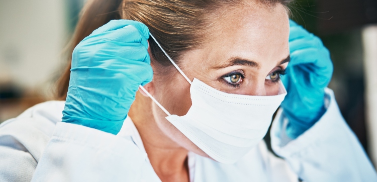 A blonde woman wearing a lab coat and surgical gloves puts on a surgical mask.